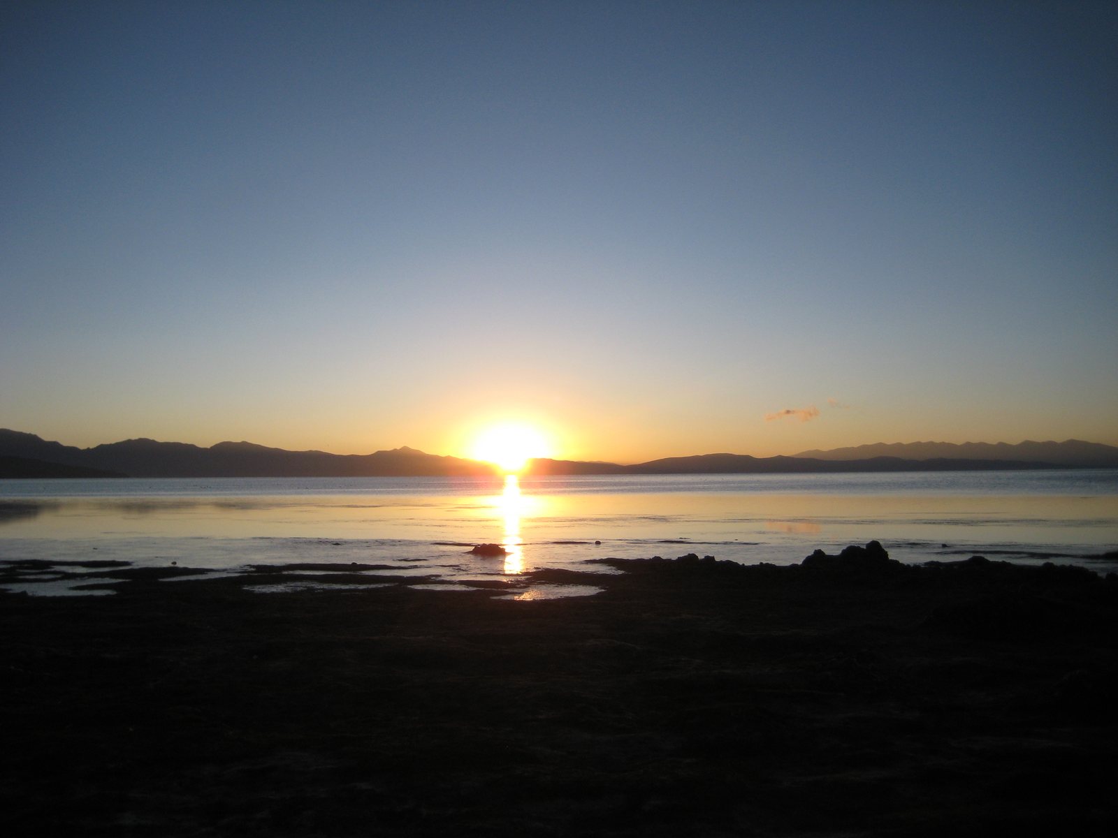 Die tief stehende Sonne über einem weiten, ruhigen See, dahinter eine Bergkette in Silhouette
